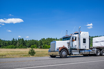 Fototapeta na wymiar Powerful big rig American semi truck tractor transporting covered fastened cargo on flat bed semi trailer driving on the highway summer road at sunny day