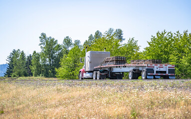 Fototapeta premium Powerful bonnet big rig beige semi truck transporting fastened by slings lumber wood on the flat bed semi trailer driving on the road in scenic nature area