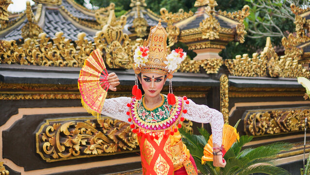 girl wearing Balinese traditional dress with a dancing gesture on Balinese temple background with hand-held fan, crown, jewelry, and gold ornament accessories. Balinese dancer woman portrait
