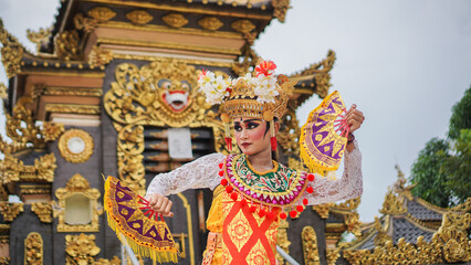 girl wearing Balinese traditional dress with a dancing gesture on Balinese temple background with hand-held fan, crown, jewelry, and gold ornament accessories. Balinese dancer woman portrait
