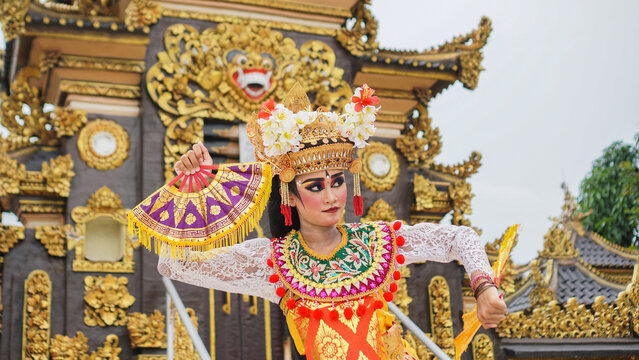 girl wearing Balinese traditional dress with a dancing gesture on Balinese temple background with hand-held fan, crown, jewelry, and gold ornament accessories. Balinese dancer woman portrait
