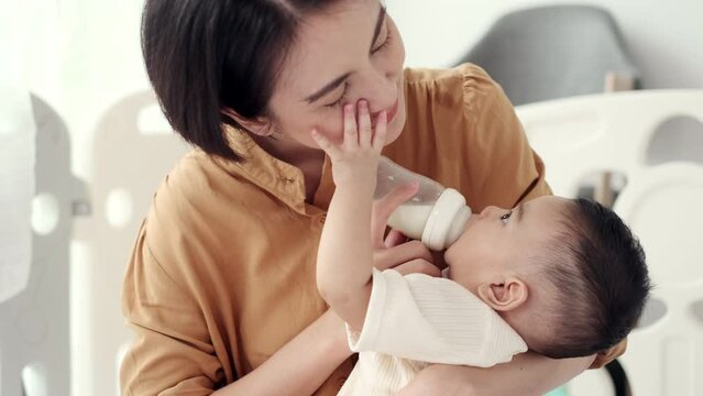 Happy young Asian mother feeding cute baby boy with milk from baby bottle. Little baby drinking milk and touching on face mother. mothers day concept.