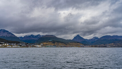 From the side of the Beagle Canal, a coastal strip with city houses is visible. A picturesque mountain range of the Andes against a cloudy sky. Ripples on the water. Ushuaia. Argentina.