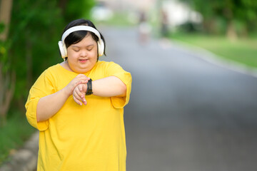 Asian girl with down syndrome wearing headphones and looking at smartwatch smiling happily walking...