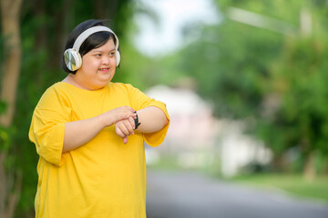 Asian girl with down syndrome wearing headphones and looking at smartwatch smiling happily walking...