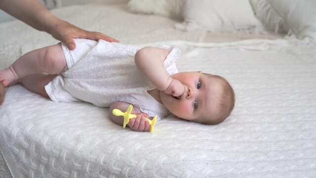 An 8-month-old baby is lying on a bed on a white blanket with a teether in his hand in a relaxed position. Mother's hand stroking the baby. First teeth. High quality 4k footage