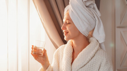 Obraz na płótnie Canvas Morning hydration. Water balance. Spa therapy. Relaxed satisfied woman with clean face in bathrobe towel enjoying drinking from glass at light home interior with free space.