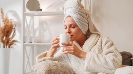 Morning coffee. Weekend relaxation. Spa leisure. Happy inspired woman in bathrobe with mug enjoying hot drink aroma on couch after bath at light home interior with free space. - obrazy, fototapety, plakaty