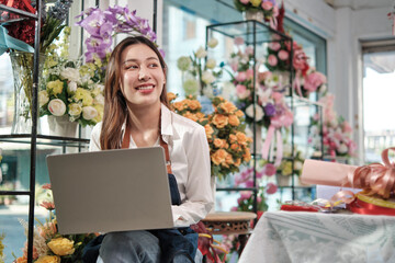 One young Asian female florist and SME entrepreneur in apron, works with laptop, online selling floral arrange in colorful flower shop store with beautiful bunch of blossoms, and e-commerce business.