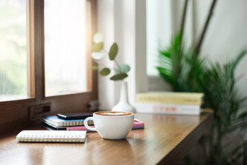 Close-up view, white coffee cup on wooden counter and smart phone, note book put on digital tablet by window, coffee break and relax after work in cafe with a reading book in the background