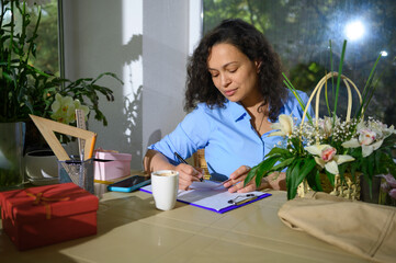 Beautiful woman writing on clipboard, working in cozy small floral shop. Flowwer arrangements and gifts concept