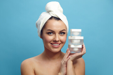 Cute smiling young woman in a head towel holding various three moisturizer jars for skin near her face on a blue isolated background. Beauty, spa, skin care.