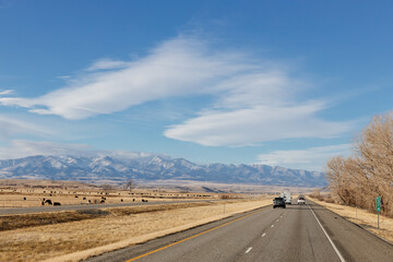 Landscape with a highway among meadows and trees in the foreground, blue mountains with snow in the background and a bright blue sky with large white clouds. Scenic landscape on a sunny autumn day