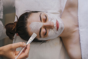 Top view of crop anonymous female applying facial mask on face while lying on bed with closed eyes