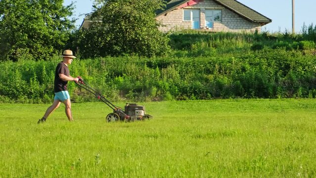 A man is mowing a lawn with a lawn mower in green summer garden. A professional gardener with a lawnmower cares for the grass, side view. Lawn cultivation. Large field. It is sunny day. Trimming work.