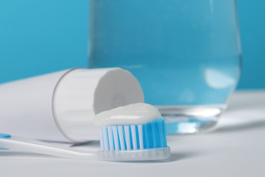 Plastic toothbrush with paste and glass of water on white table against light blue background, closeup