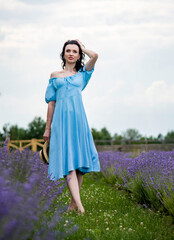 Naklejka na ściany i meble beautiful brunette smiling girl in blue dress walking in the blooming field of lavender flowers and holding straw hat in her hands