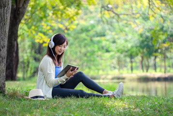young attractive asian woman sitting in spring park,smiling,wearing headphones to listen music on online tablet i the morning sunshine,concept people lifestyle,holiday,relax,health benefit in nature