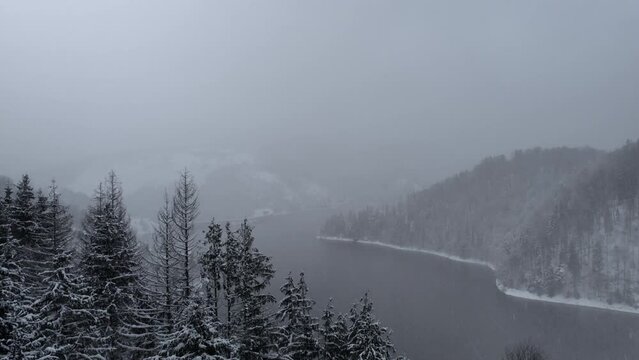 Aerial panorama of a lake and dam surrounded by a captivating blend of fog and snow, mountain forest, aerial lifting up,in Dragan dam from Transylvania, Romania