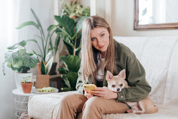 Freelance woman with mobile phone and dog, and working from home office. Happy girl sitting on couch in living room with plants. Distance learning and online education.