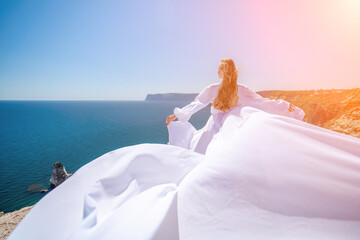 woman sea white dress. Blonde with long hair on a sunny seashore in a white flowing dress, rear view, silk fabric waving in the wind. Against the backdrop of the blue sky and mountains on the seashore