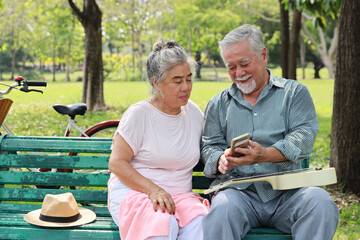 Happy smiling asian senior man and woman sitting on bench and using smartphone with ukulele in garden park outdoor. Musical and relaxation makes lover couple happiness. Health care lifestyle concept.
