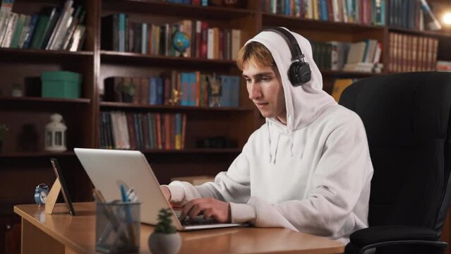 The guy is sitting at the table, obviously playing a computer game. He has a hood on his head and headphones on top of it. He is highly focused on the game.