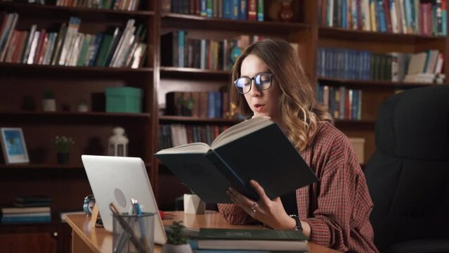 The woman is reading a book while talking to someone on a webcam, showing the book she is reading. She is sitting in a chair, and there are many books on a shelf in the room.