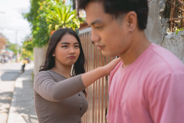 An asian lady wearing a brown long sleeves top is trying to console the guy in pick shirt by putting her hand in his shoulder, looking at him sincerely and acting cute by pouting.