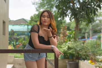 A pretty lady wearing a black blouse and jeans shorts is leaning on a rail, posing with an open palm gesture, tight lip and a squinted eyes towards the camera. Plants and trees in the background.