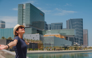 travel to the United Arab Emirates, Happy young asian female traveler in dress and hat look at View of Abu Dhabi Skyline in Al Maryah Island. Vacation and tourist destination concept.