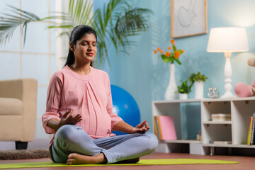 Indian peaceful pregnant woman in lotus pose doing meditation on yoga mat home - concept of mindfulness, relaxation and Self-care during pregnancy - obrazy, fototapety, plakaty