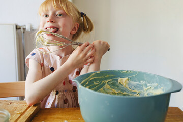 Happy girl tasting cake batter from wire whisk at home
