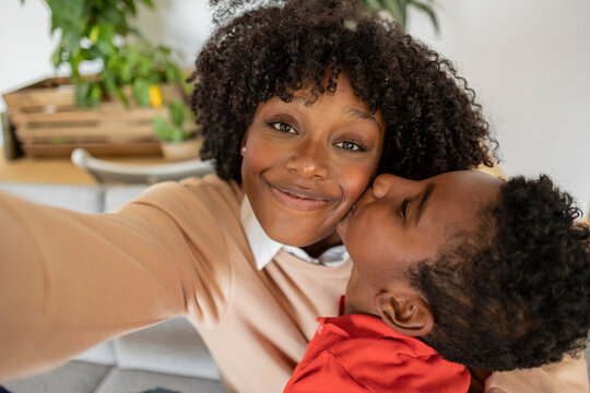 Smiling woman taking selfie with boy kissing on cheek at home