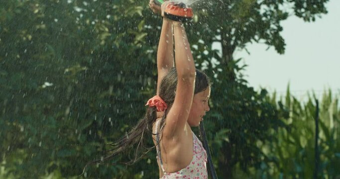 Slow Motion Portrait of Carefree Happy Little Girl in Swimsuit Playing with Water and Having Fun in Garden. Female Child Splashing Herself with Water Sprinkler on Sunny Summer Day, Enjoying Childhood