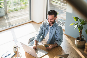 Happy businessman sitting with smart phone and using laptop at home office