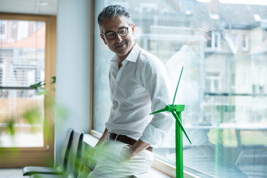 Smiling businessman looking at wind turbine model leaning on window sill in office