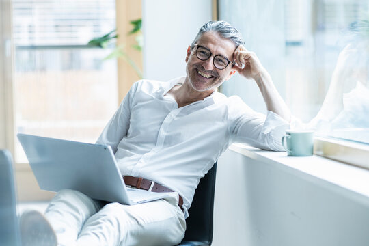 Happy businessman with laptop leaning head on window sill at office