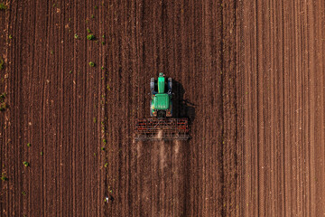Green tractor vehicle with tiller attached performing field tillage before the sowing season, aerial shot seen from the drone pov