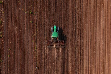 Poster Green tractor vehicle with tiller attached performing field tillage before the sowing season, aerial shot seen from the drone pov © Bits and Splits