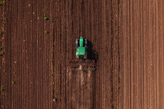 Fototapeta Green tractor vehicle with tiller attached performing field tillage before the sowing season, aerial shot seen from the drone pov