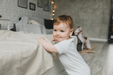 Fototapeta premium A cute little baby 6-9 months old is standing near the bed.