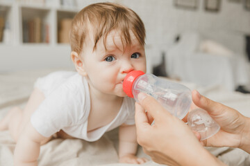 A baby with big blue eyes on a white bed with a special bottle of water with a pacifier. Tries to chew on it. teething