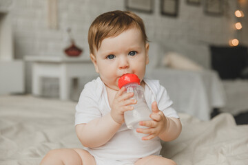 A baby with big blue eyes on a white bed with a special bottle of water with a pacifier. Tries to chew on it. teething