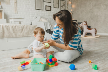Naklejka premium Beautiful young mother plays and teaches her baby 6 months old on the floor in the living room.mom and baby play with toys