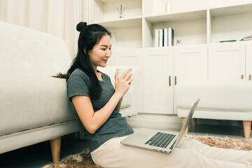 Asian woman is sitting in the living room holding a coffee cup and a laptop computer on her lap at home.