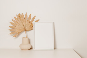 Empty aesthetic photo frame mockup with vase and palm leaf on wooden white table. Picture frame...