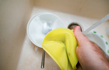 A person holds an yellow silicone sponge above a sink full of sparkling clean water, washing dishes with perfect POV.