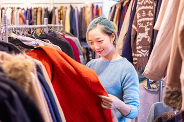 Stylish Asian woman is choosing second hand sweater from the clothing shop for winter season...