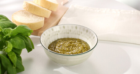 pesto sauce in bowl and on bread. basil and slices of bread on a white tablecloth. traditional italian green sauce. angle view. sunny day.